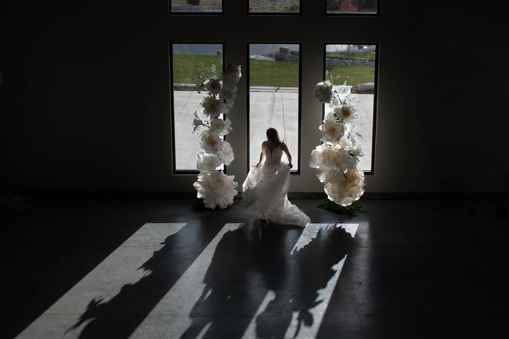 From a higher vantage point, a bride holds her lace gown’s train while standing before floor-to-ceiling windows and giant white paper flowers, her shadow cast on the concrete floor.