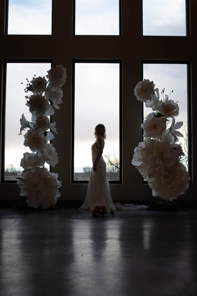 Silhouetted bride in a detailed lace gown stands in front of expansive windows, framed by oversized white floral decor on either side. A bride, seen in profile against the light, poses between two towering white floral installations and tall windows, creating a dramatic silhouette.