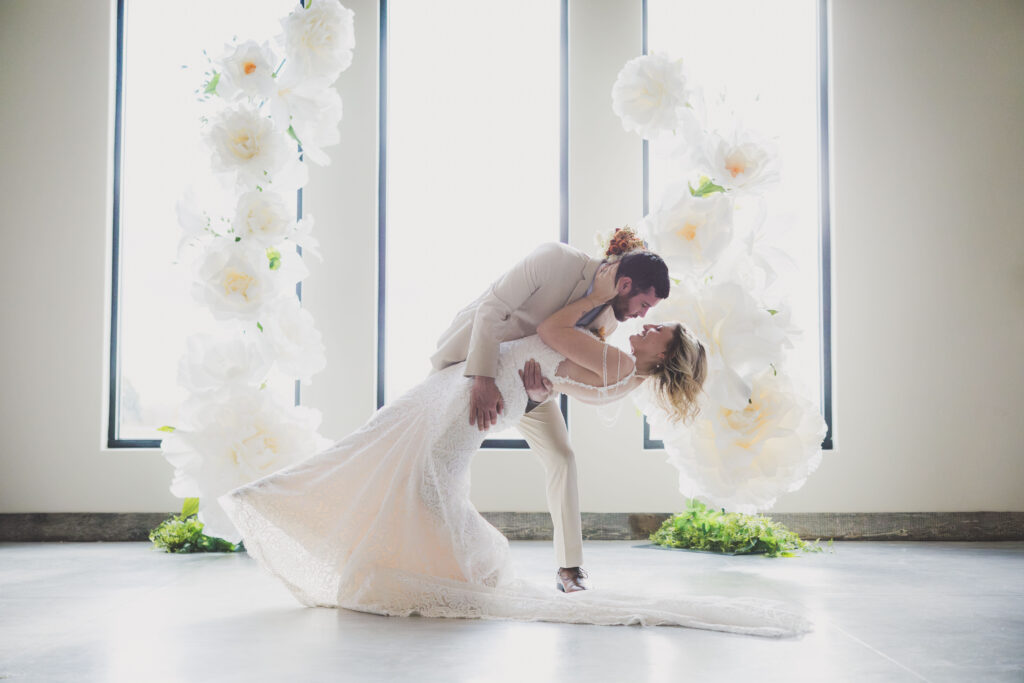 A bride in a lace gown is dipped backward by her groom in a light tan suit in front of tall windows, surrounded by large white floral arrangements.